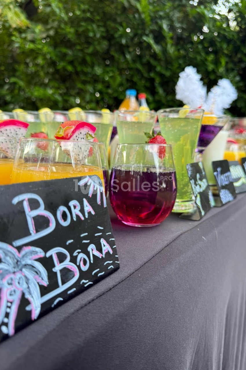 Verres de cocktails colorés alignés sur une table avec des ardoises décorées, en extérieur près de végétation.