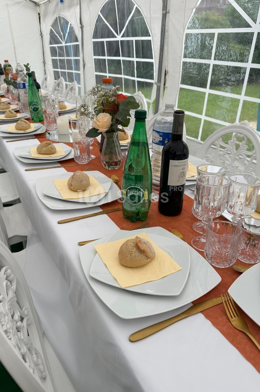 Table dressée sous une tente avec assiettes, verres, pains, bouteilles d'eau et de vin, décorée de fleurs.