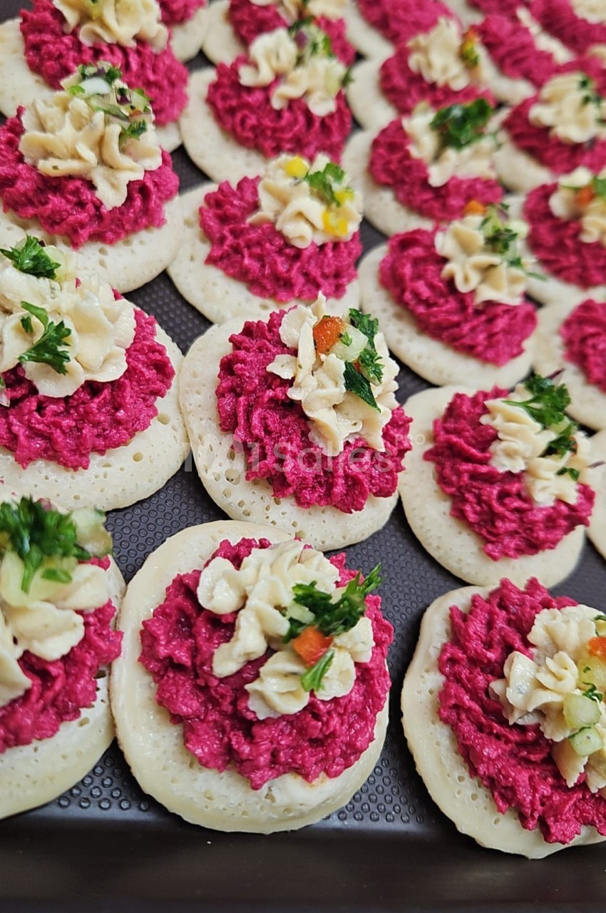 Plateau de mini-blinis garnis de mousse rose, crème décorative et herbes fraîches.