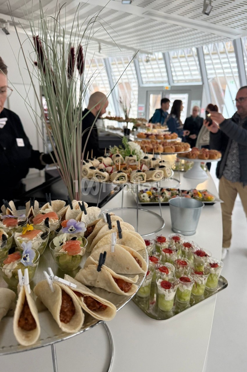 Buffet varié avec wraps, verrines et amuse-bouches, présenté dans une salle lumineuse avec des convives en arrière-plan.