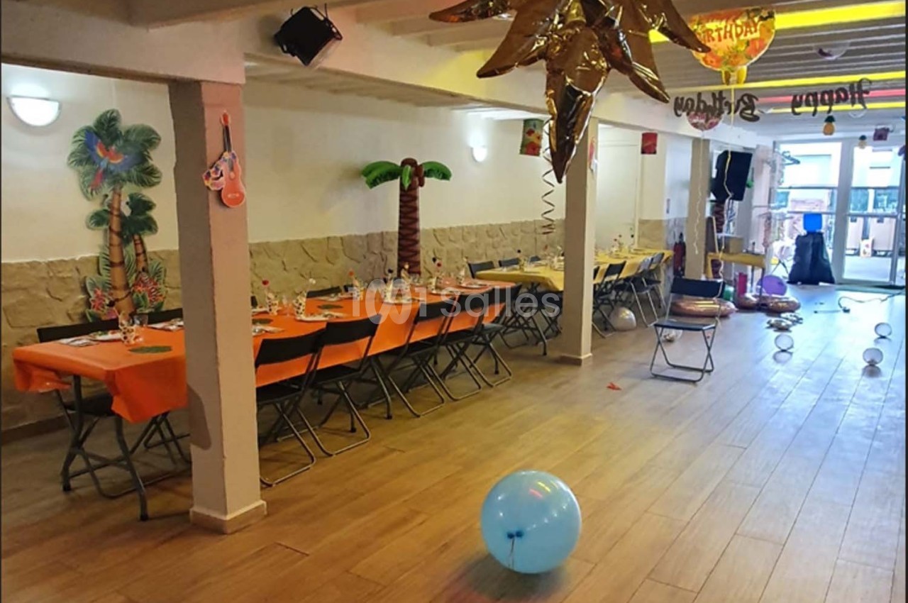 Salle décorée pour une fête avec tables dressées, ballons colorés et ornements tropicaux.