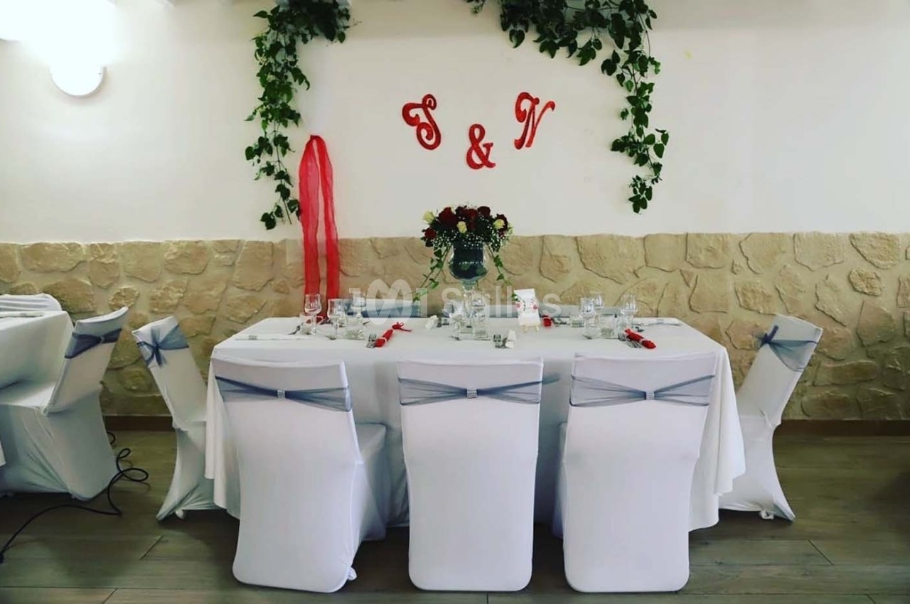 Table décorée pour un événement avec nappes blanches, chaises ornées de rubans, fleurs au centre et initiales murales.