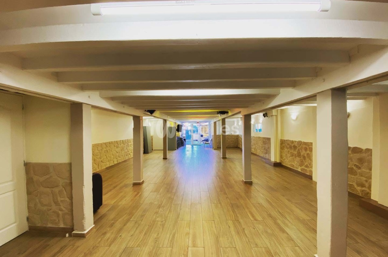 Salle vide avec sol en parquet, murs clairs partiellement recouverts de pierre, et poutres blanches au plafond.