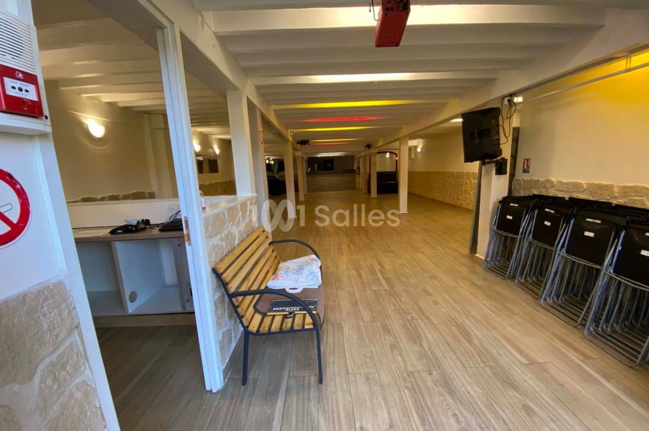 Salle vide avec sol en bois, murs clairs, rangées de chaises empilées et un banc en bois près de l'entrée.