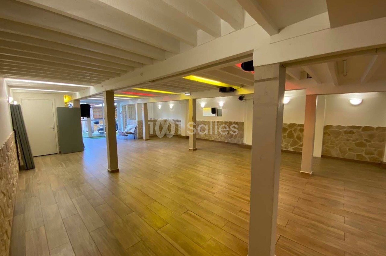 Salle spacieuse avec sol en parquet, murs en pierre et poutres blanches apparentes, éclairée par des lumières modernes.