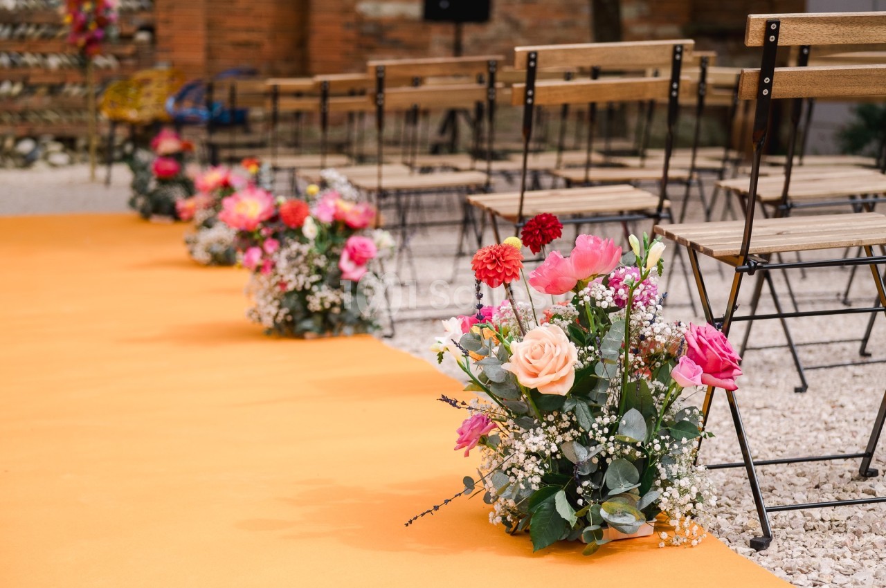 Allée décorée de fleurs colorées et de chaises en bois, disposées pour une cérémonie en extérieur.