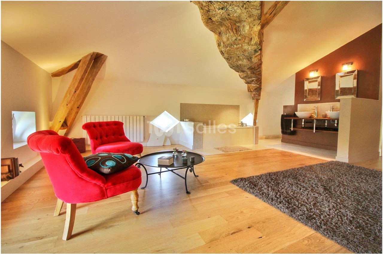 Salon mansardé avec poutres apparentes, fauteuils rouges, table basse, parquet en bois clair et salle de bain attenante.