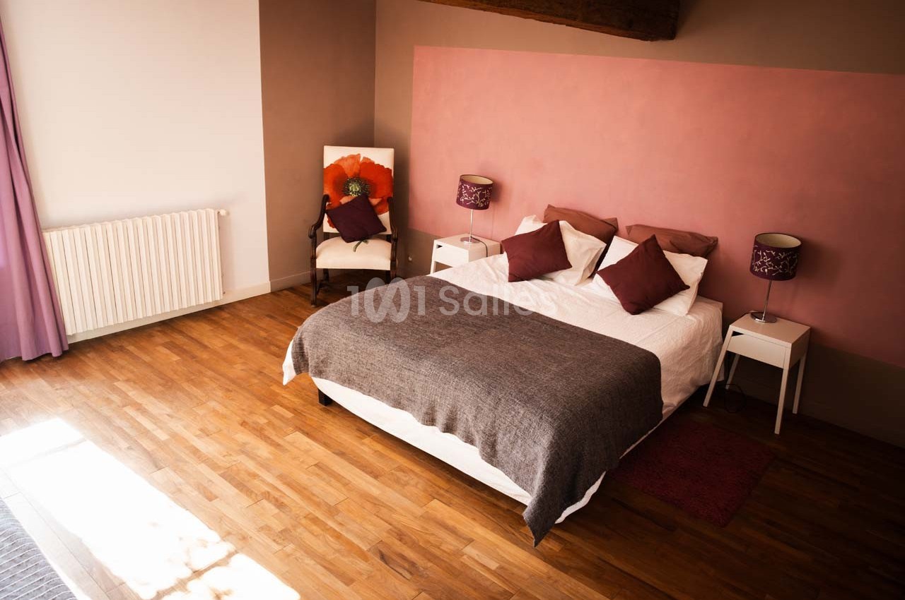 Chambre avec lit double, couvre-lit gris, coussins rouges, murs rose et beige, parquet et fauteuil avec coussin orange.