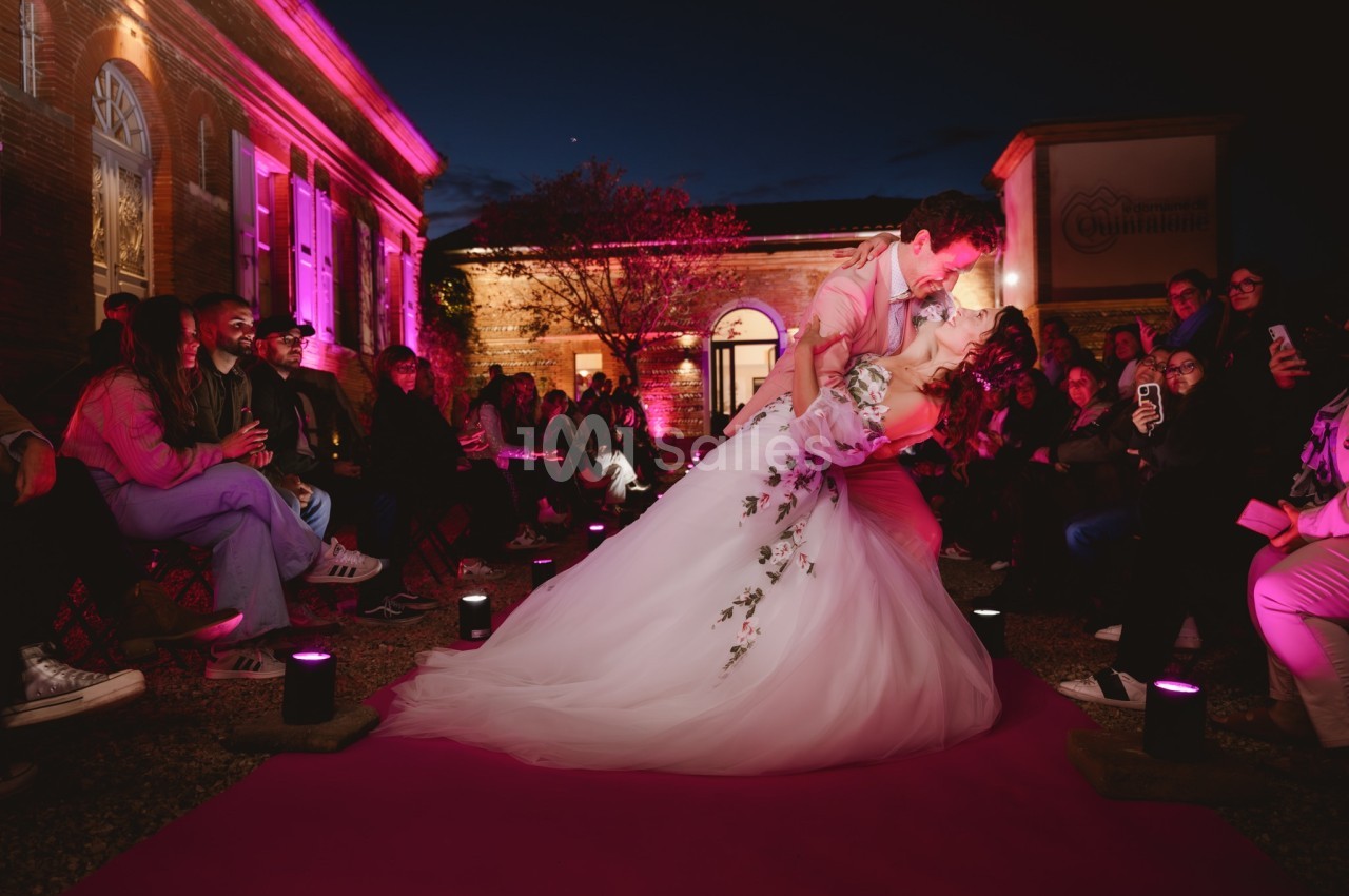 Un couple de mariés danse sur un tapis rose, entouré d'invités assis, dans une ambiance nocturne éclairée en rose.