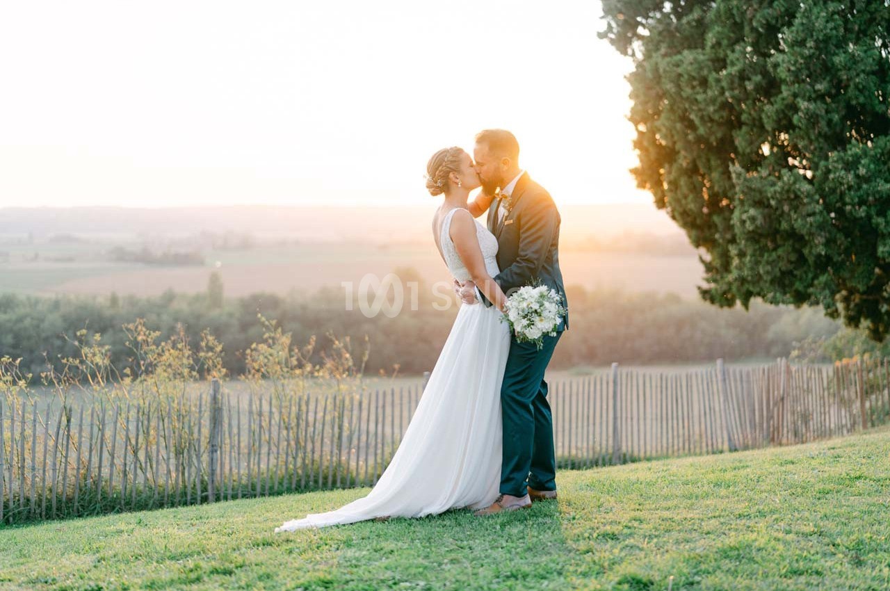 Un couple en tenue de mariage s'embrasse dans un jardin au coucher du soleil, près d'une clôture et d'un arbre.