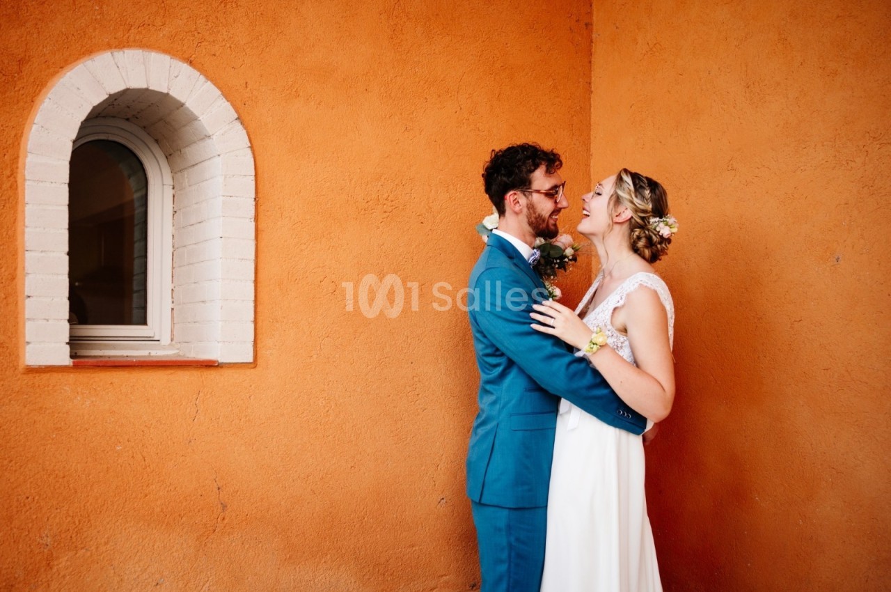 Un couple en tenue de mariage s'enlace devant un mur orange avec une petite fenêtre voûtée.