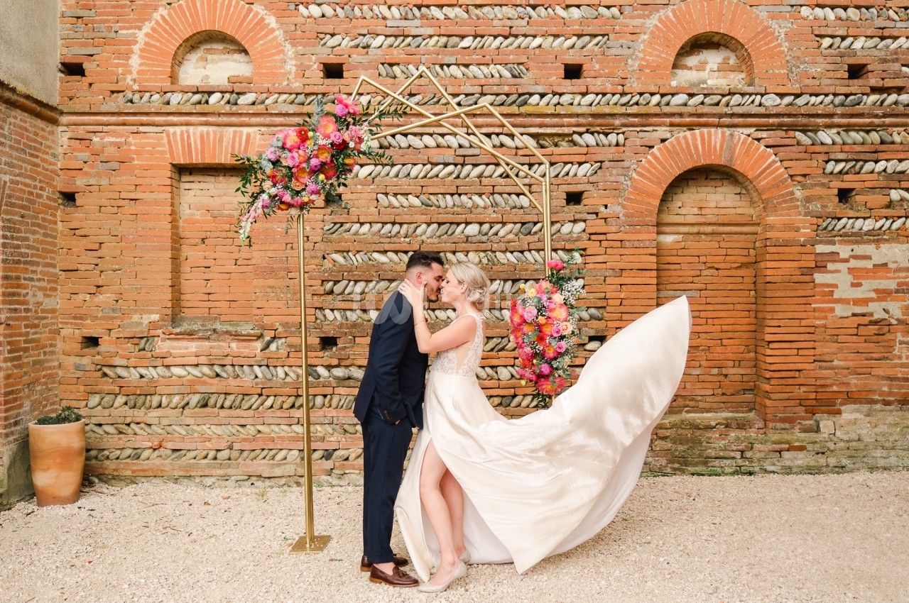 Un couple de mariés s'embrasse sous une arche décorée de fleurs colorées, devant un mur en briques anciennes.