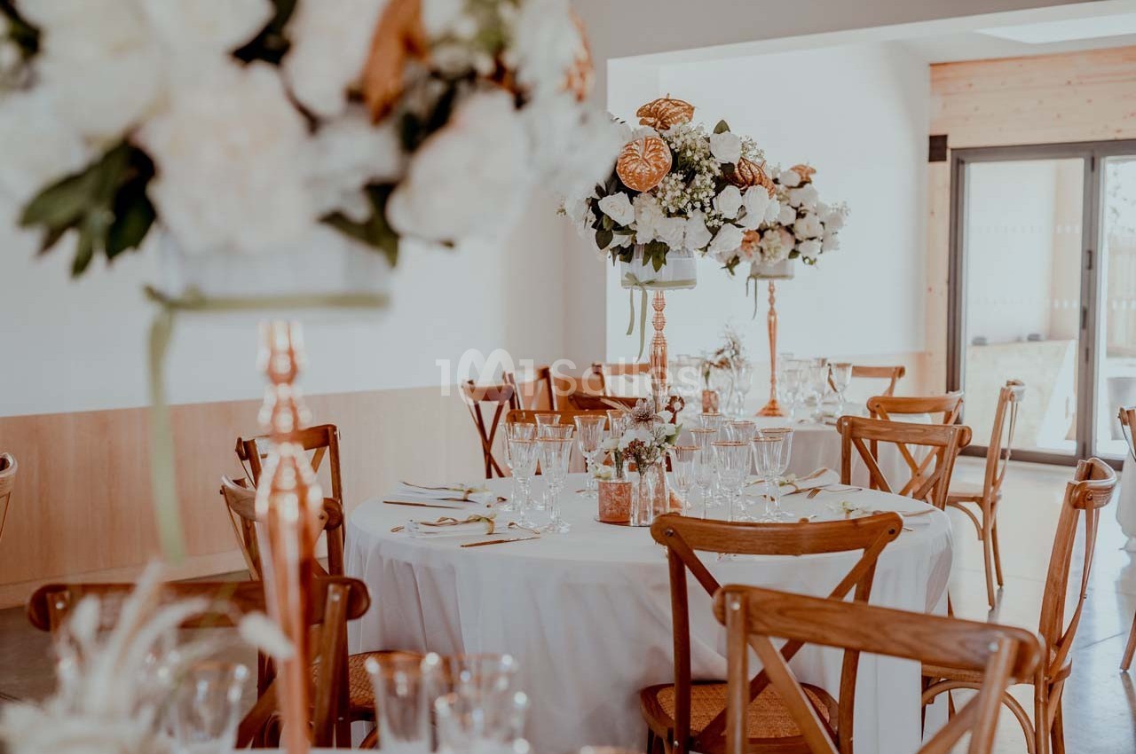 Salle décorée pour un événement avec tables rondes, nappes blanches, chaises en bois et centres de table fleuris.