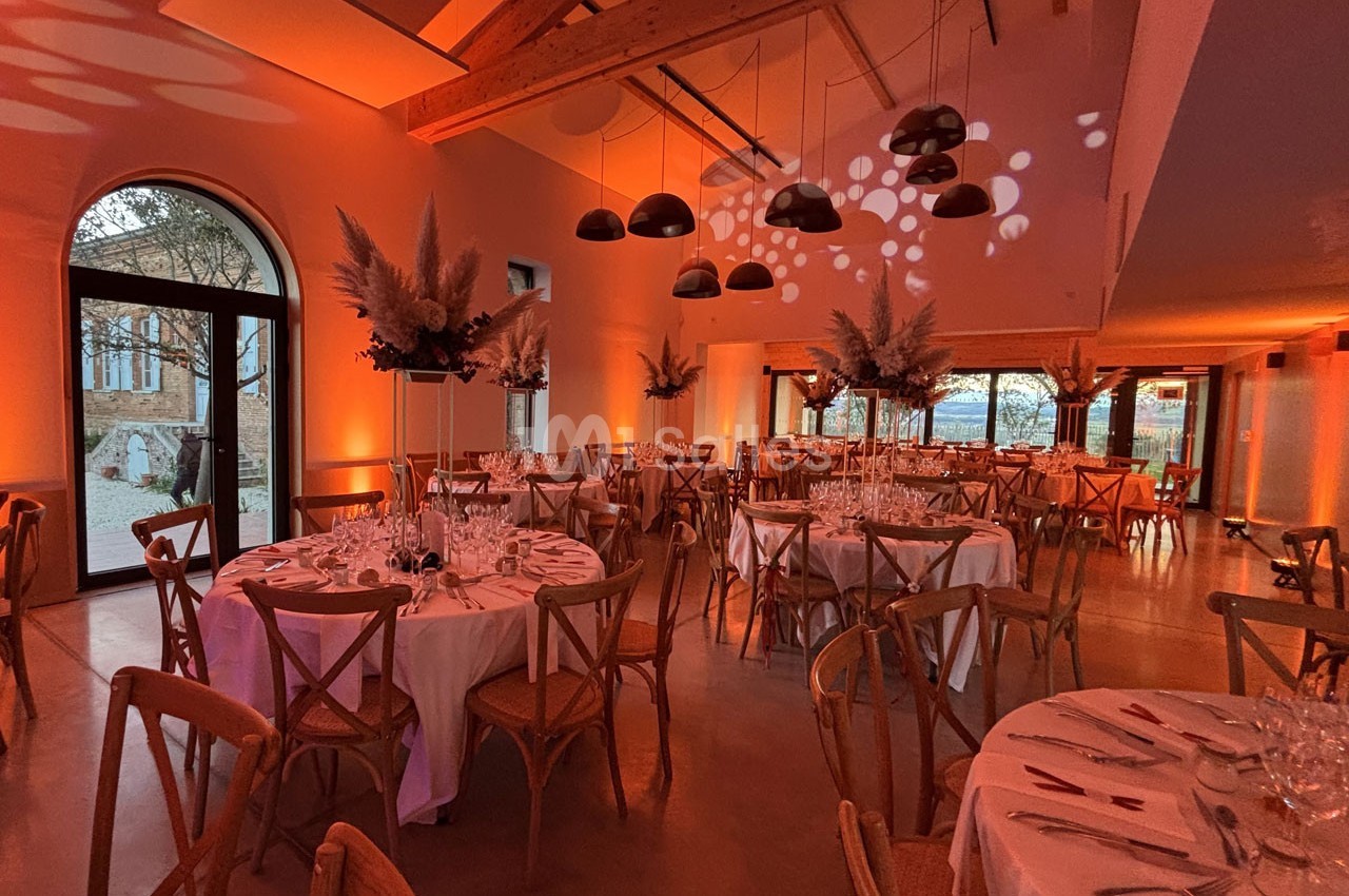 Salle de réception décorée avec des tables rondes, des chaises en bois et un éclairage tamisé aux tons chauds.