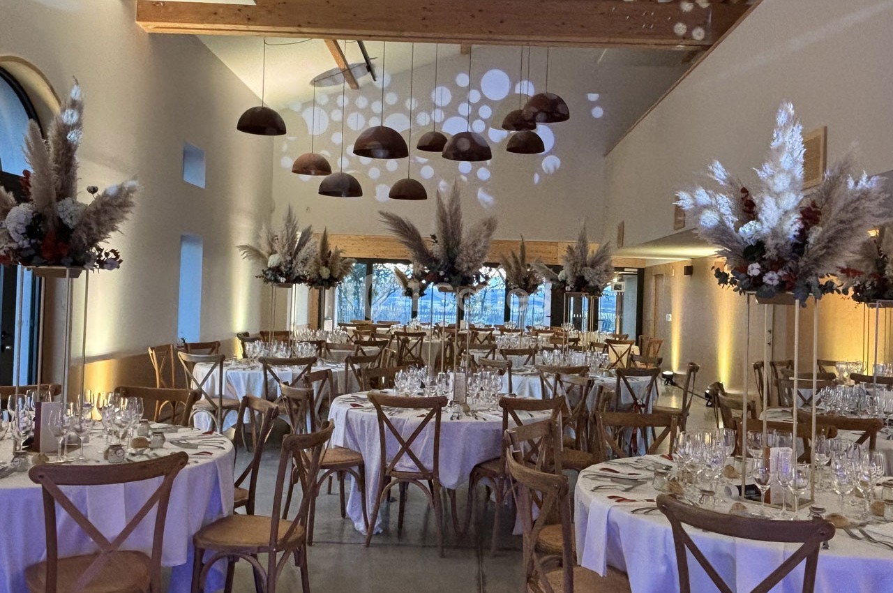 Salle de réception décorée avec des tables rondes dressées, des chaises en bois et des compositions florales suspendues.