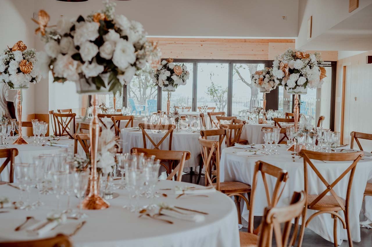 Salle de réception décorée avec des tables rondes, nappes blanches, chaises en bois et grands bouquets de fleurs.