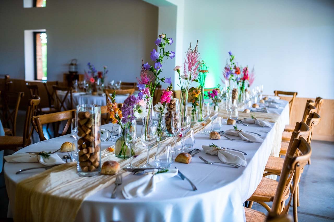 Table dressée avec nappes blanches, couverts, verres et décorations florales colorées dans une salle lumineuse.