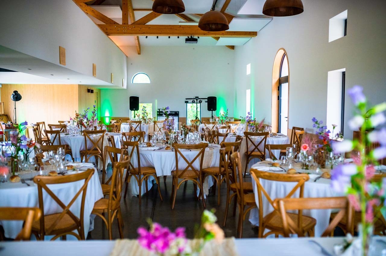 Salle de réception lumineuse avec tables rondes décorées de fleurs, chaises en bois et éclairages colorés.