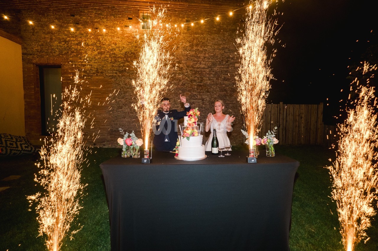 Un couple célèbre devant un gâteau de mariage entouré de fontaines d'étincelles dans un jardin en soirée.
