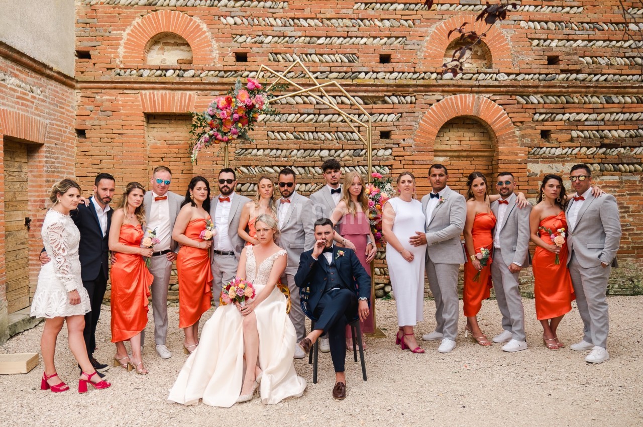 Un couple de mariés entouré de leurs invités en tenue élégante, posant devant un mur en briques décoré de fleurs.