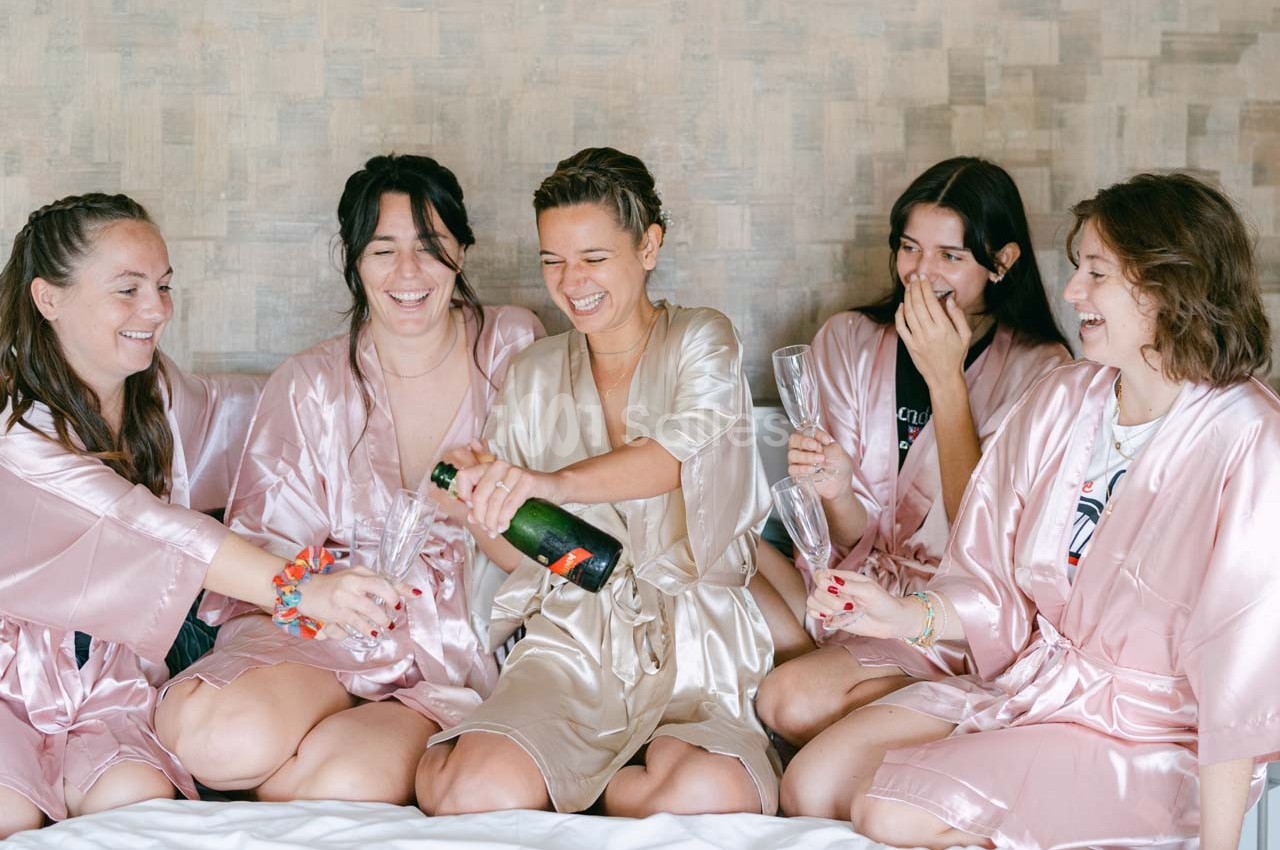 Cinq femmes en peignoirs satinés rient et trinquent avec une bouteille de champagne sur un lit.