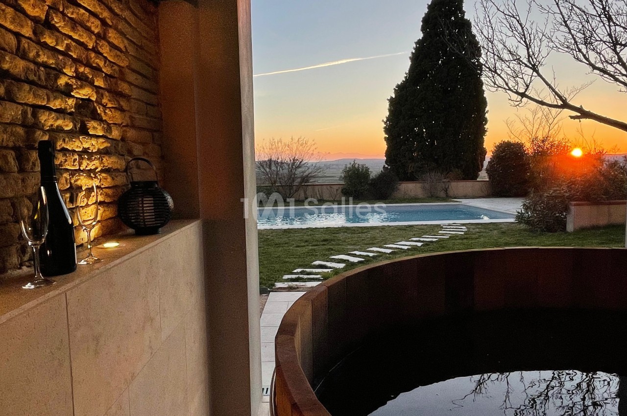 Vue d'une terrasse avec un bain nordique en bois, une piscine, un jardin et un coucher de soleil à l'horizon.