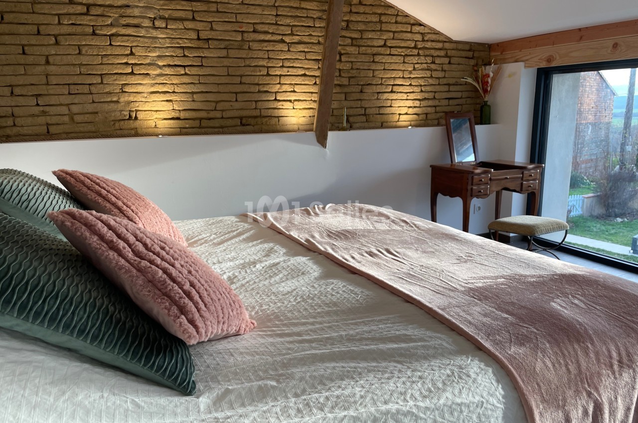 Chambre lumineuse avec un lit double, des coussins colorés, un mur en briques apparentes et un bureau près d'une fenêtre.