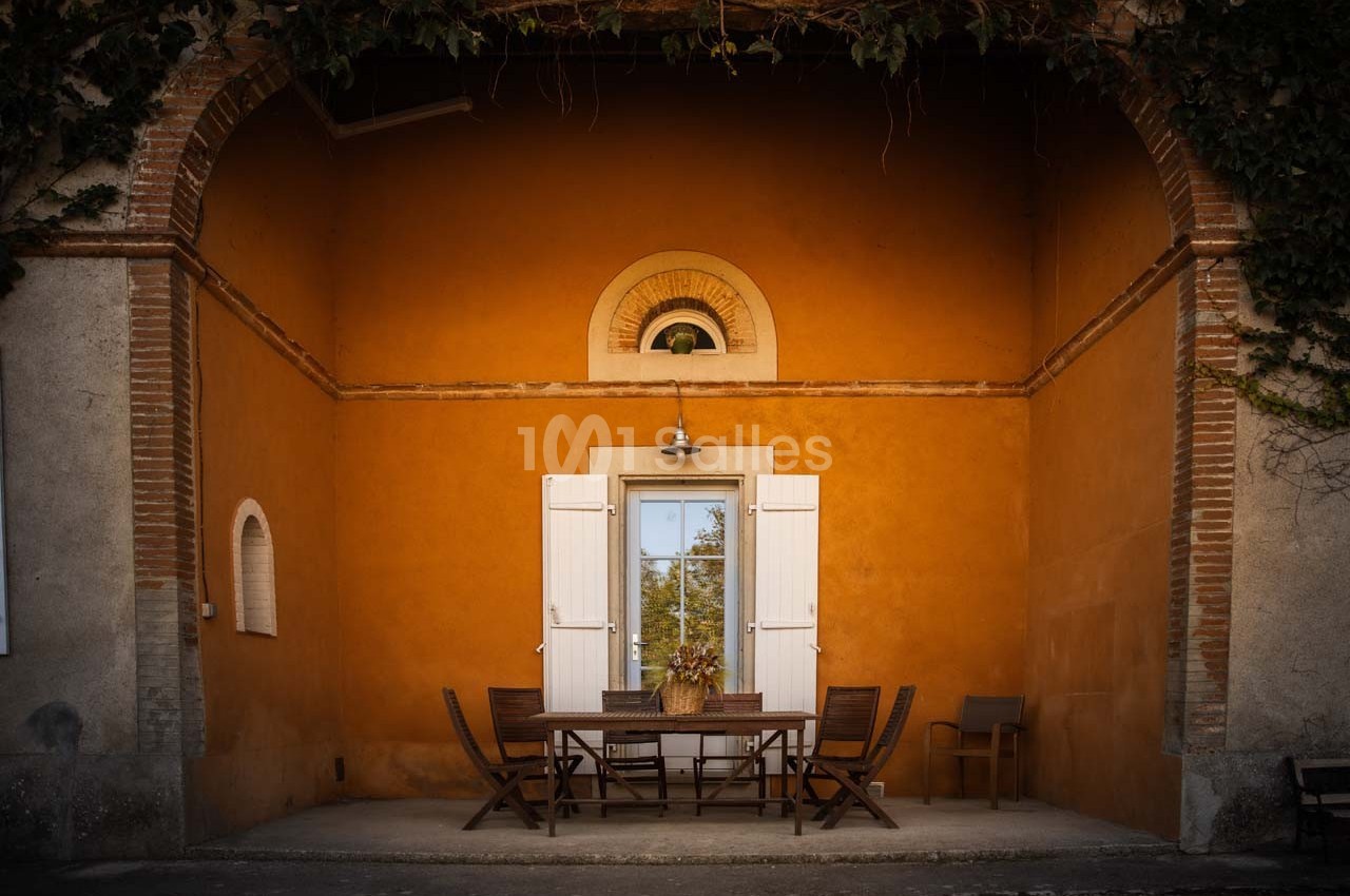 Façade orange avec une table en bois, des chaises et des volets blancs ouverts sous une arche en pierre.