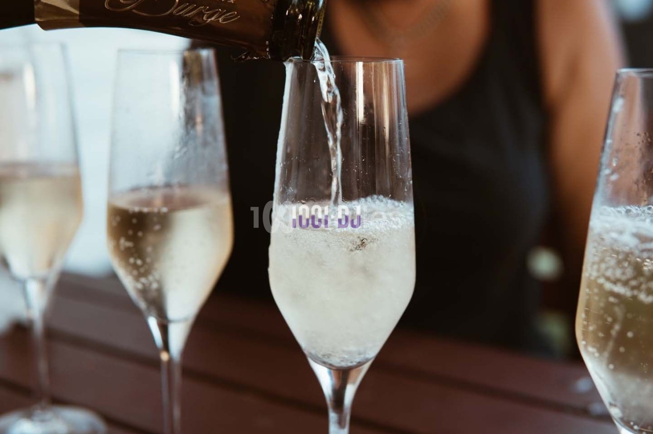 Un verre de champagne est rempli depuis une bouteille, entouré de trois autres verres sur une table en bois.