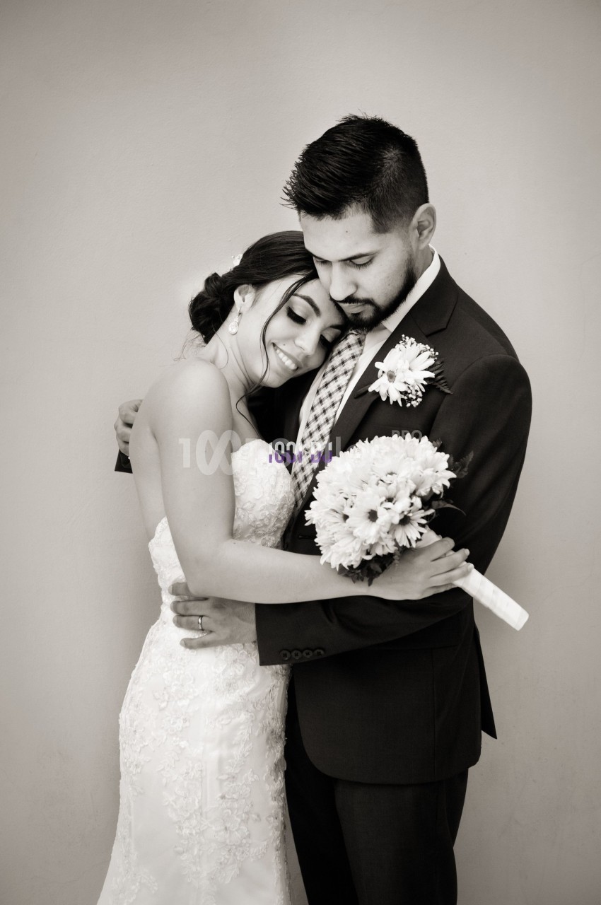 Un couple en tenue de mariage s'enlace tendrement devant un mur clair.