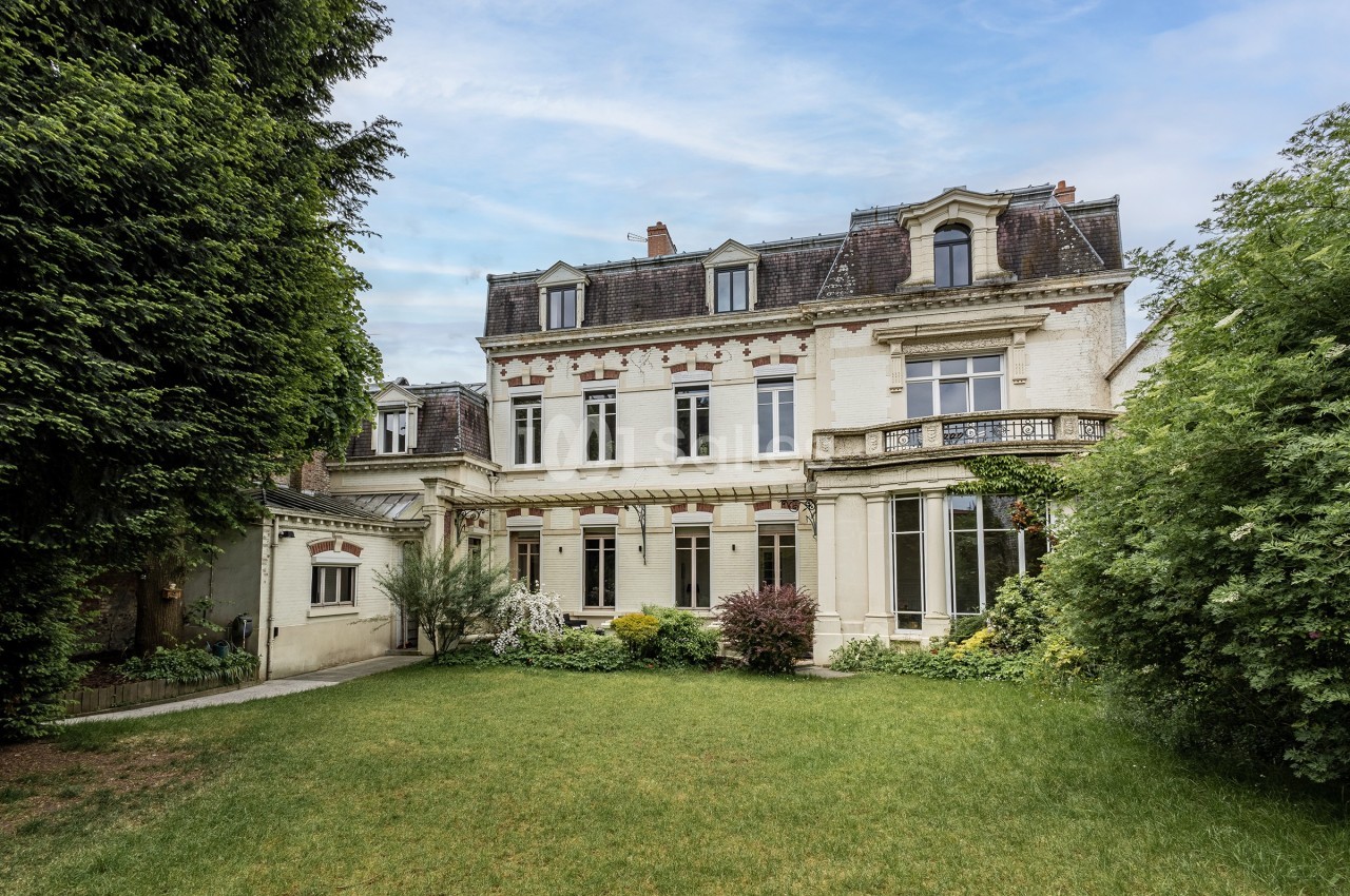 Façade arrière d'une grande maison ancienne avec jardin verdoyant et ciel dégagé en arrière-plan.