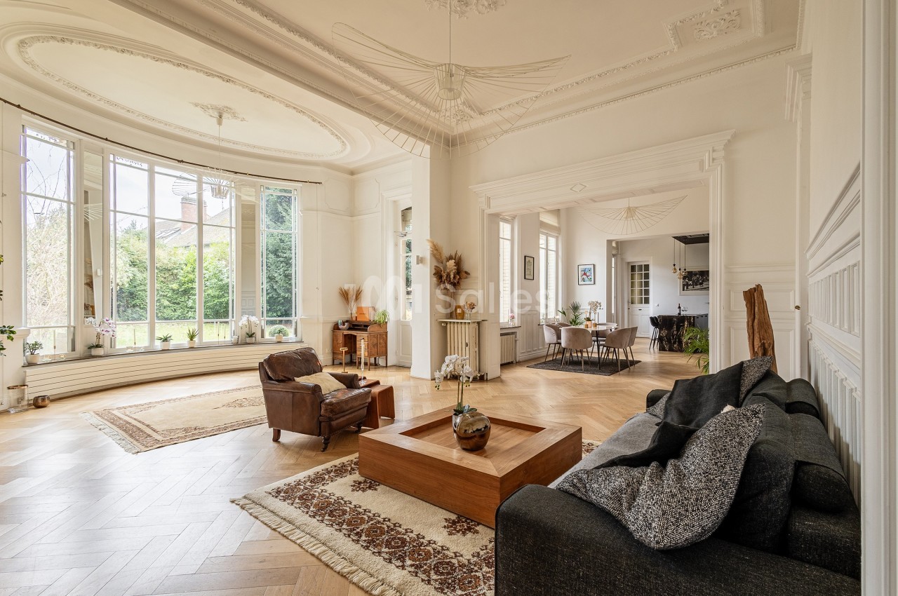 Salon lumineux avec parquet en bois, grandes fenêtres, mobilier moderne et vue sur un jardin extérieur.