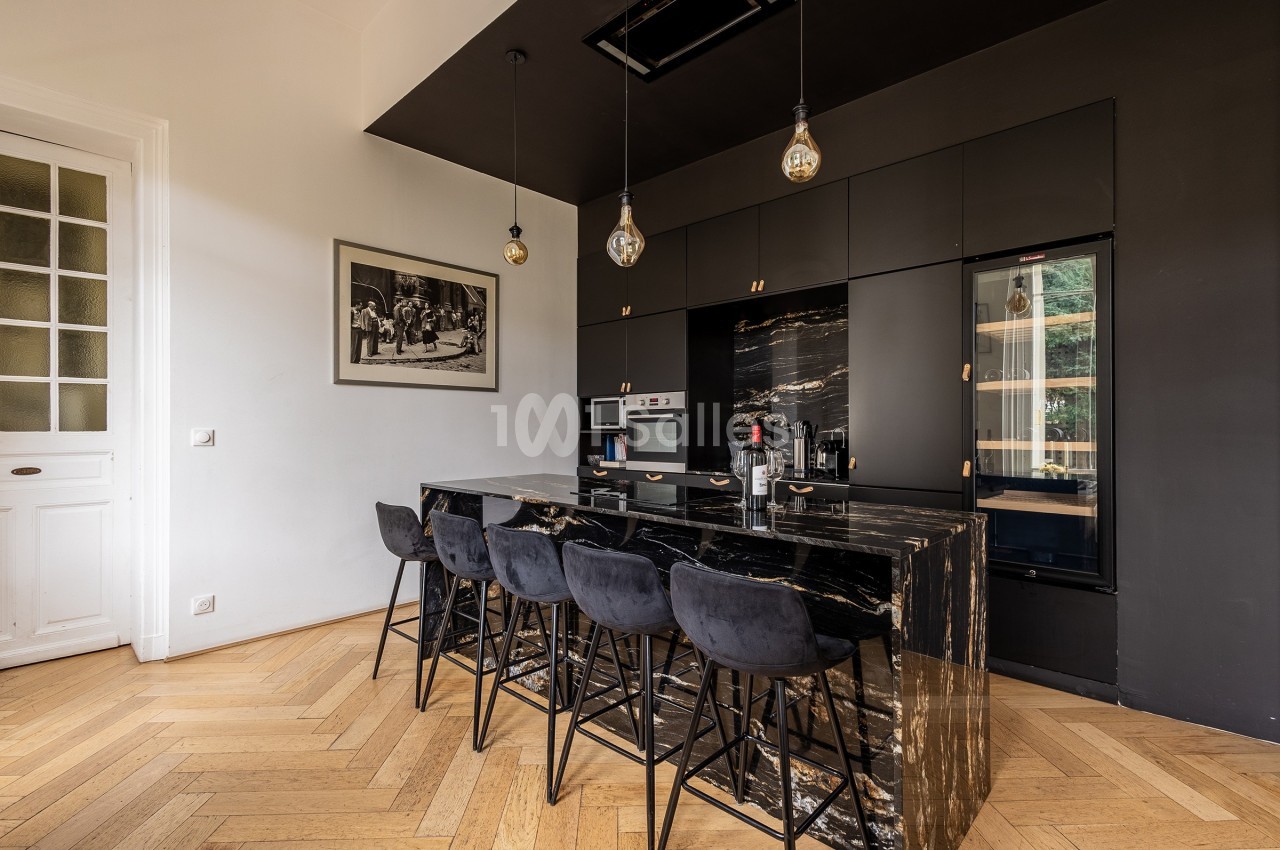 Cuisine moderne avec îlot central en marbre noir, tabourets assortis, meubles noirs et éclairage suspendu.