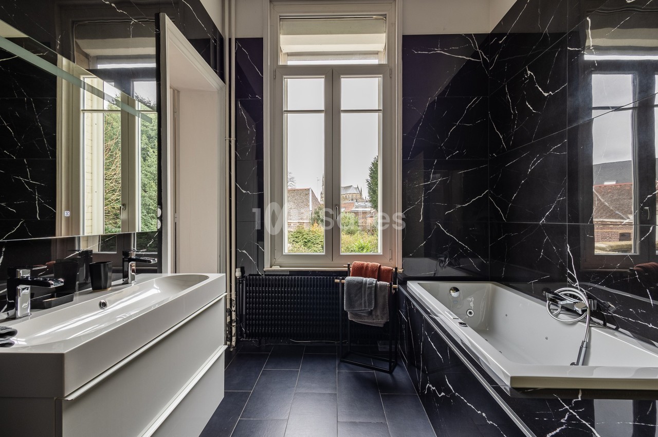 Salle de bain moderne avec murs noirs effet marbre, baignoire, lavabo blanc et grande fenêtre donnant sur un jardin.