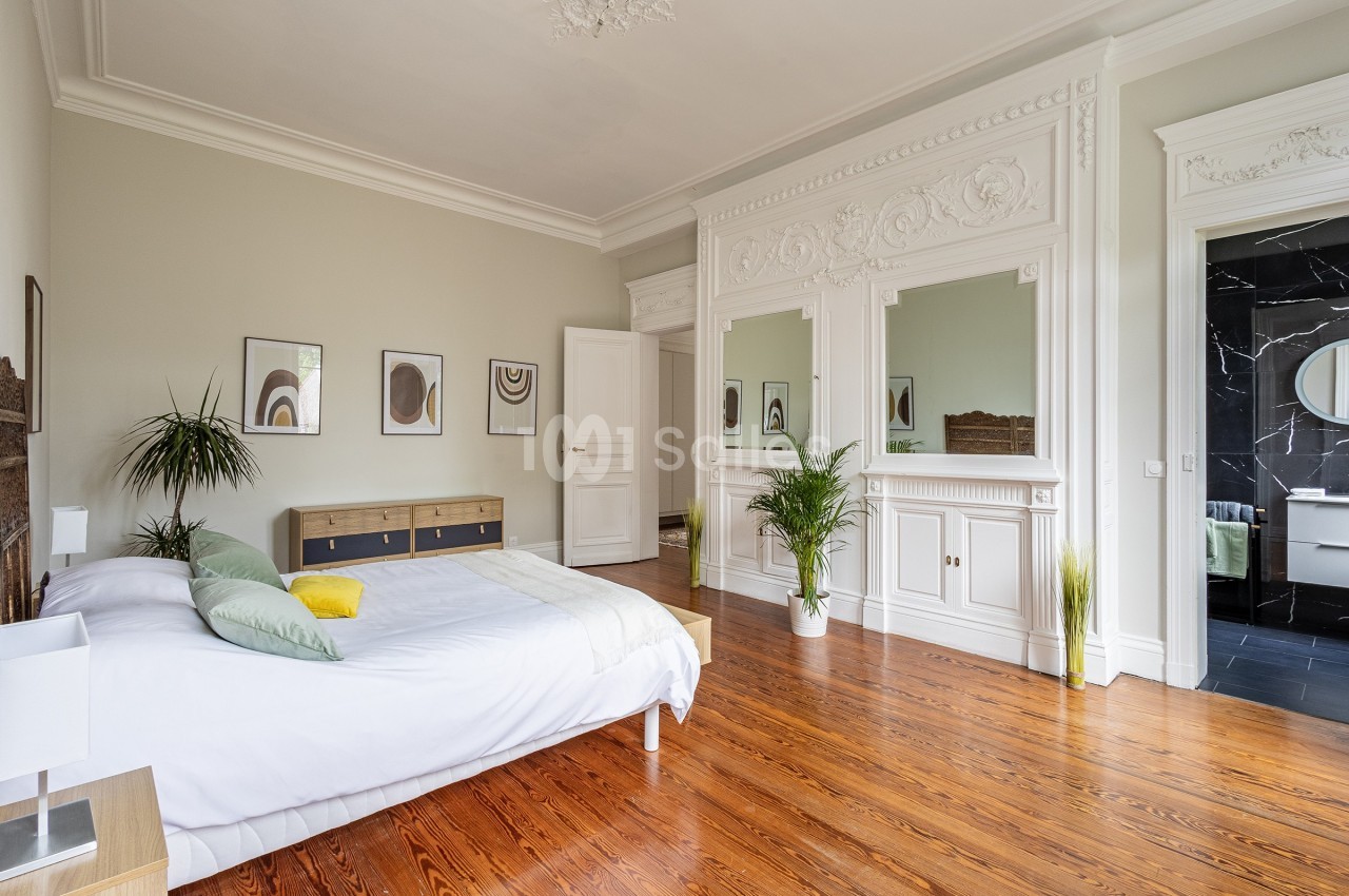 Chambre lumineuse avec parquet en bois, lit double, plantes décoratives et moulures blanches sur les murs et le plafond.
