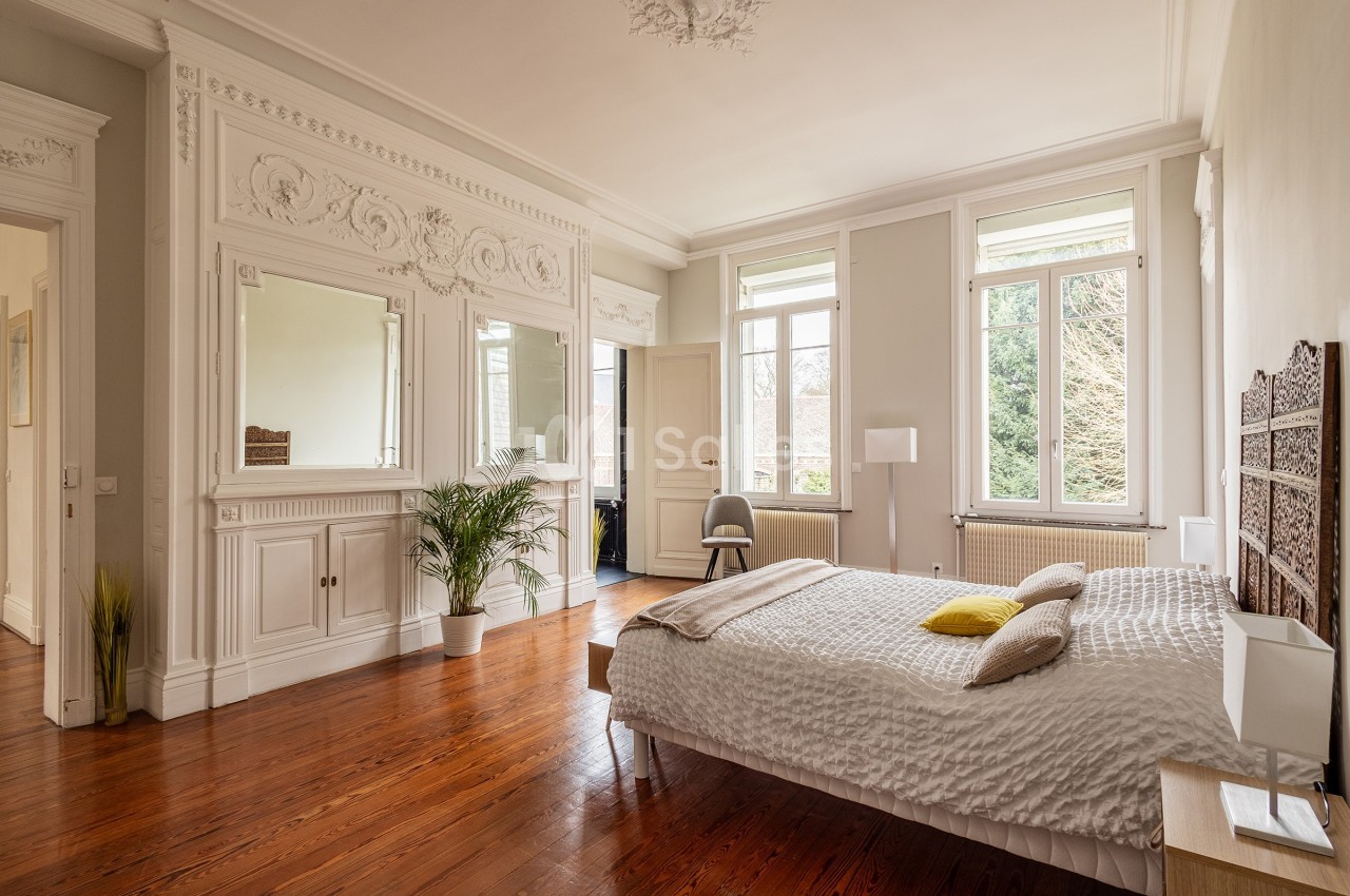 Chambre lumineuse avec parquet, lit double, moulures décoratives, miroirs et grandes fenêtres donnant sur un jardin.