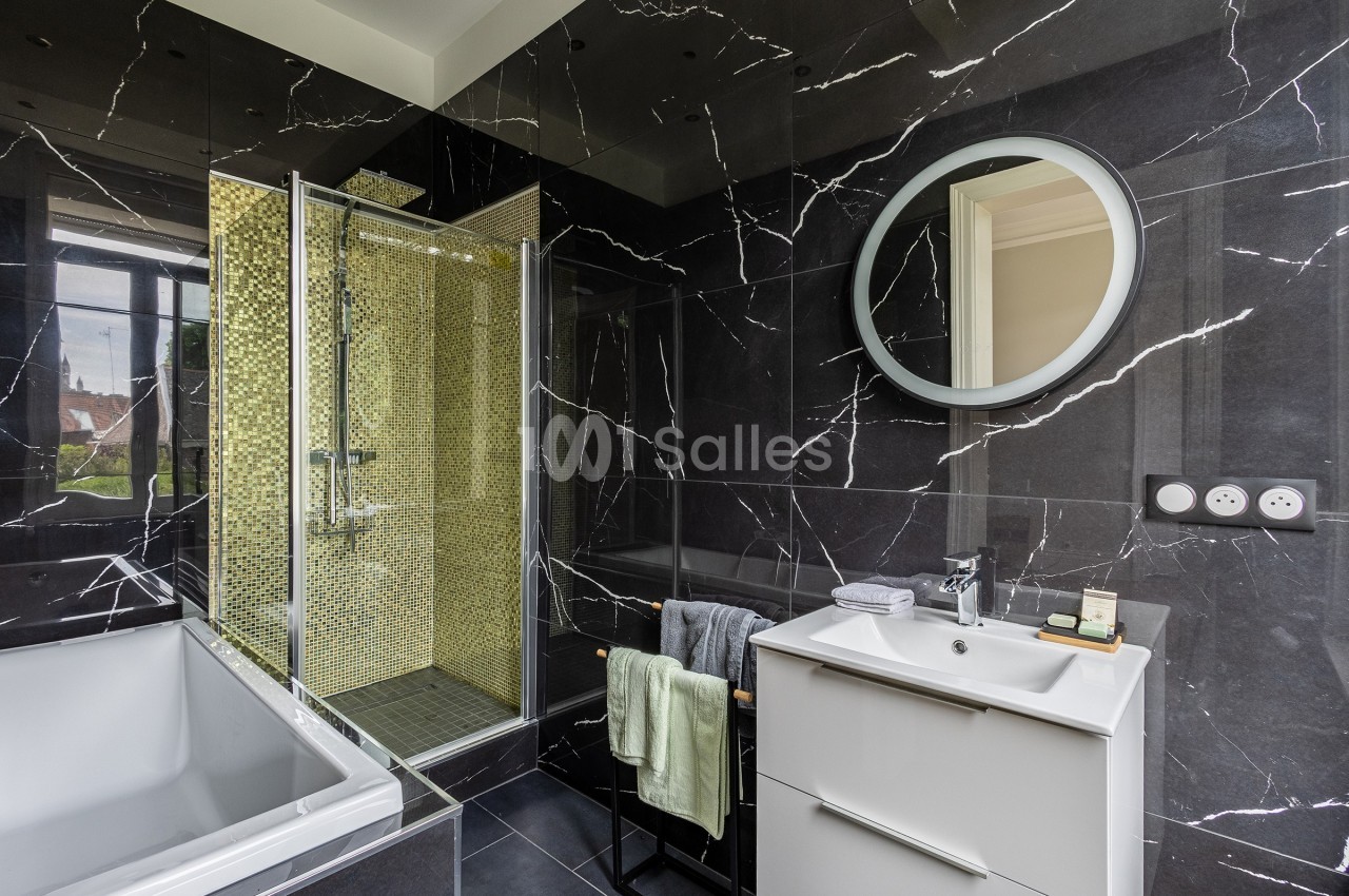 Salle de bain moderne avec murs en marbre noir, douche à mosaïque dorée, baignoire blanche et meuble vasque blanc.