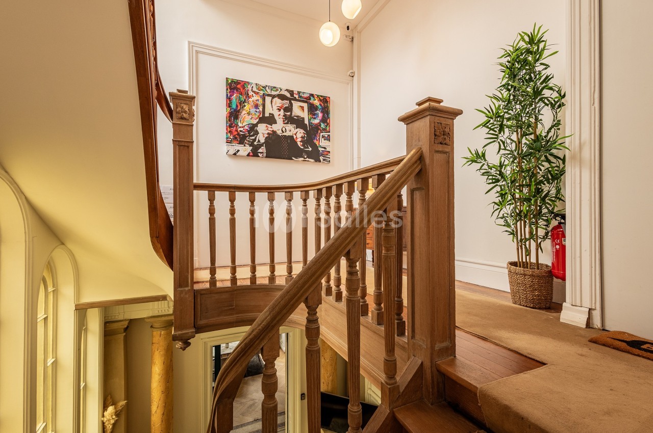 Escalier en bois avec rambarde, plante en pot et tableau coloré accroché sur un mur blanc dans un intérieur lumineux.