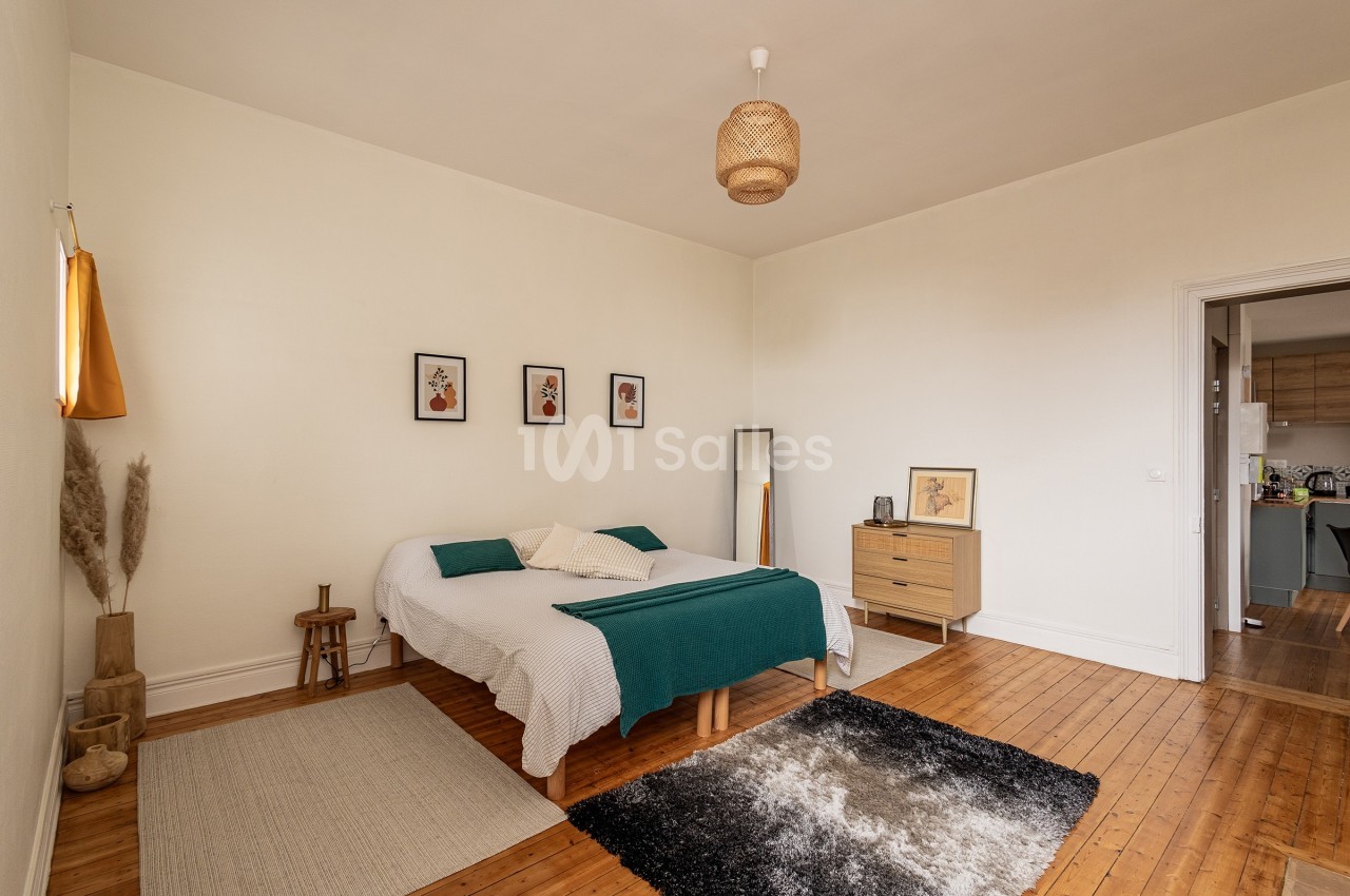 Chambre lumineuse avec lit double, décoration minimaliste, tapis au sol et vue partielle sur une cuisine adjacente.