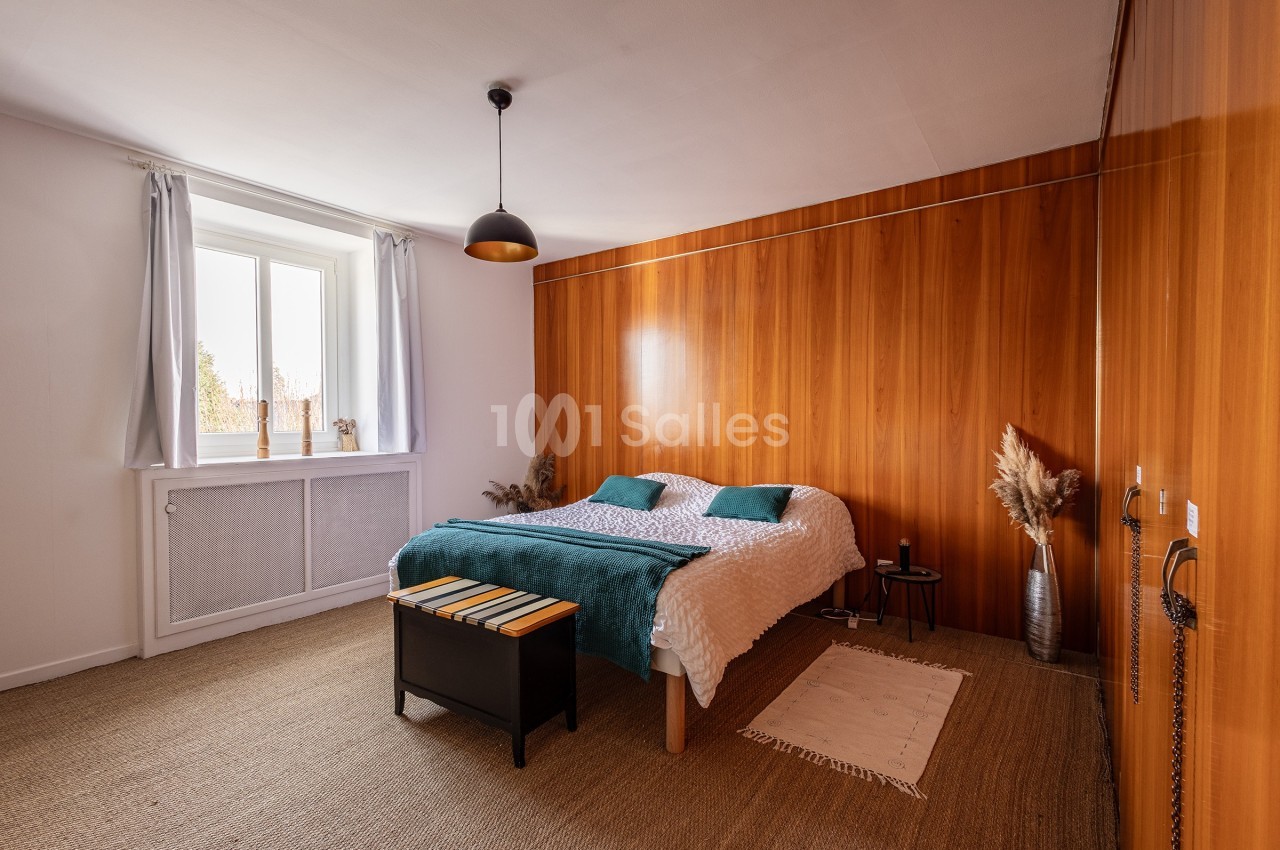 Chambre lumineuse avec lit double, mur en bois, tapis en jonc de mer, fenêtre avec rideaux blancs et décoration minimaliste.