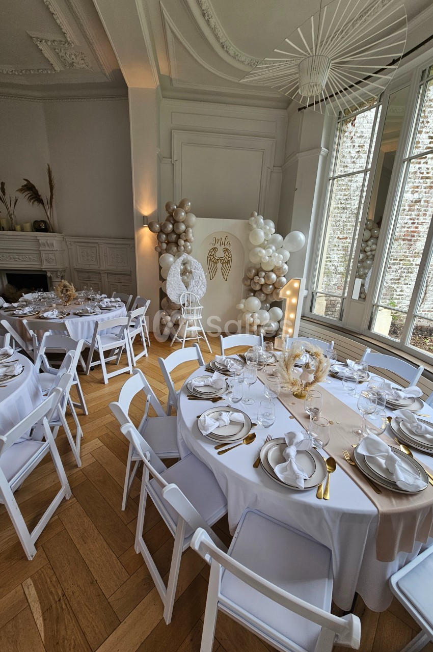 Salle décorée pour un événement avec tables dressées, nappes blanches, ballons et panneau lumineux près d'une fenêtre.
