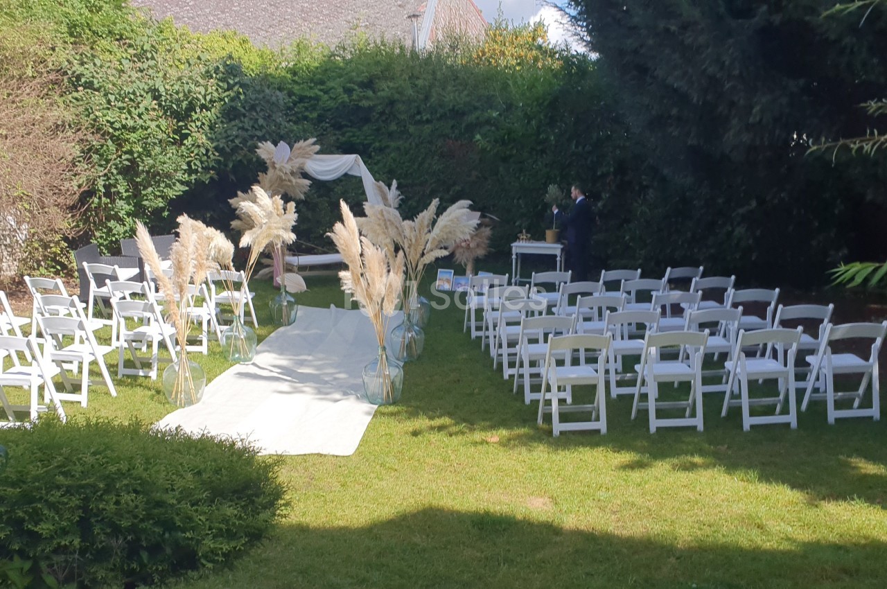 Chaises blanches disposées en extérieur autour d'une allée centrale décorée de pampas, menant à une arche de cérémonie.