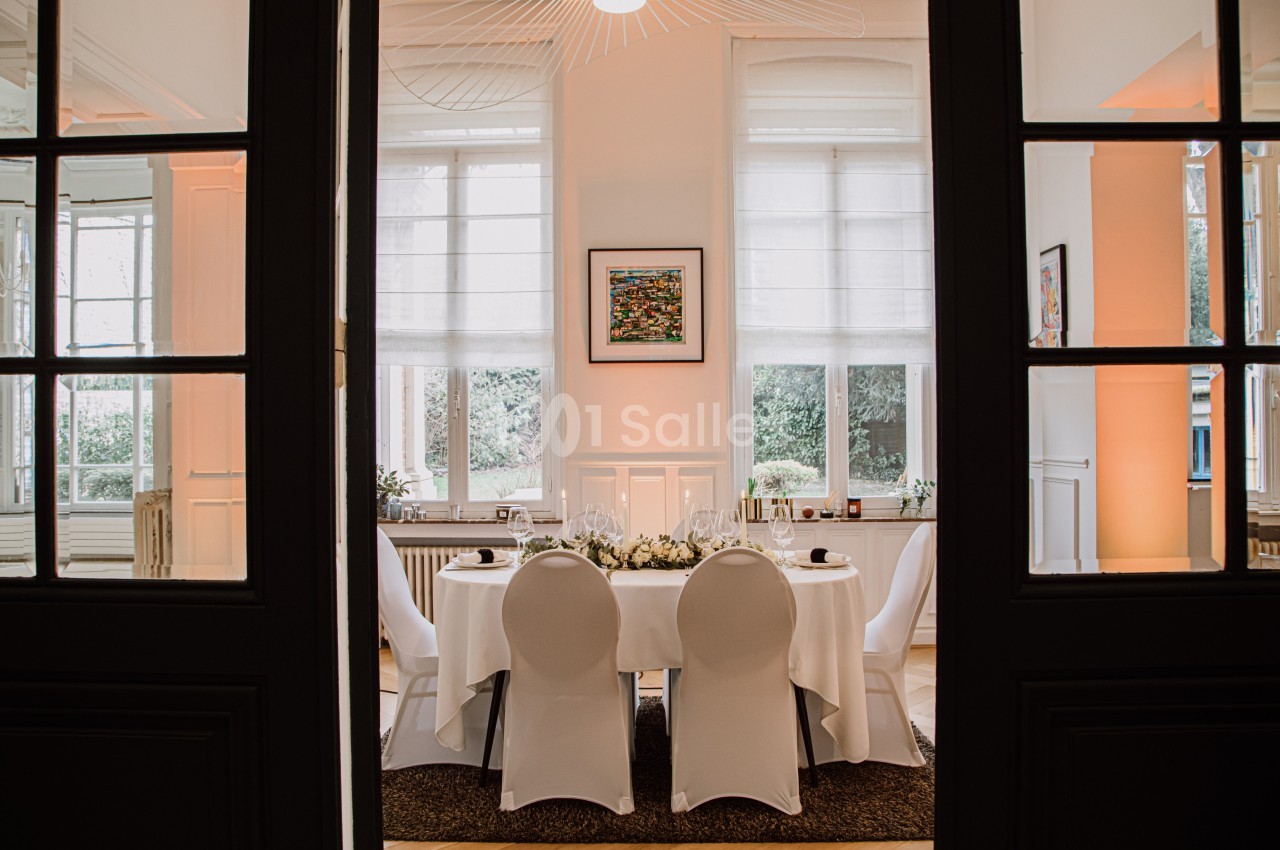 Salle élégante avec table ronde dressée pour un repas, entourée de chaises blanches et éclairée par des fenêtres.