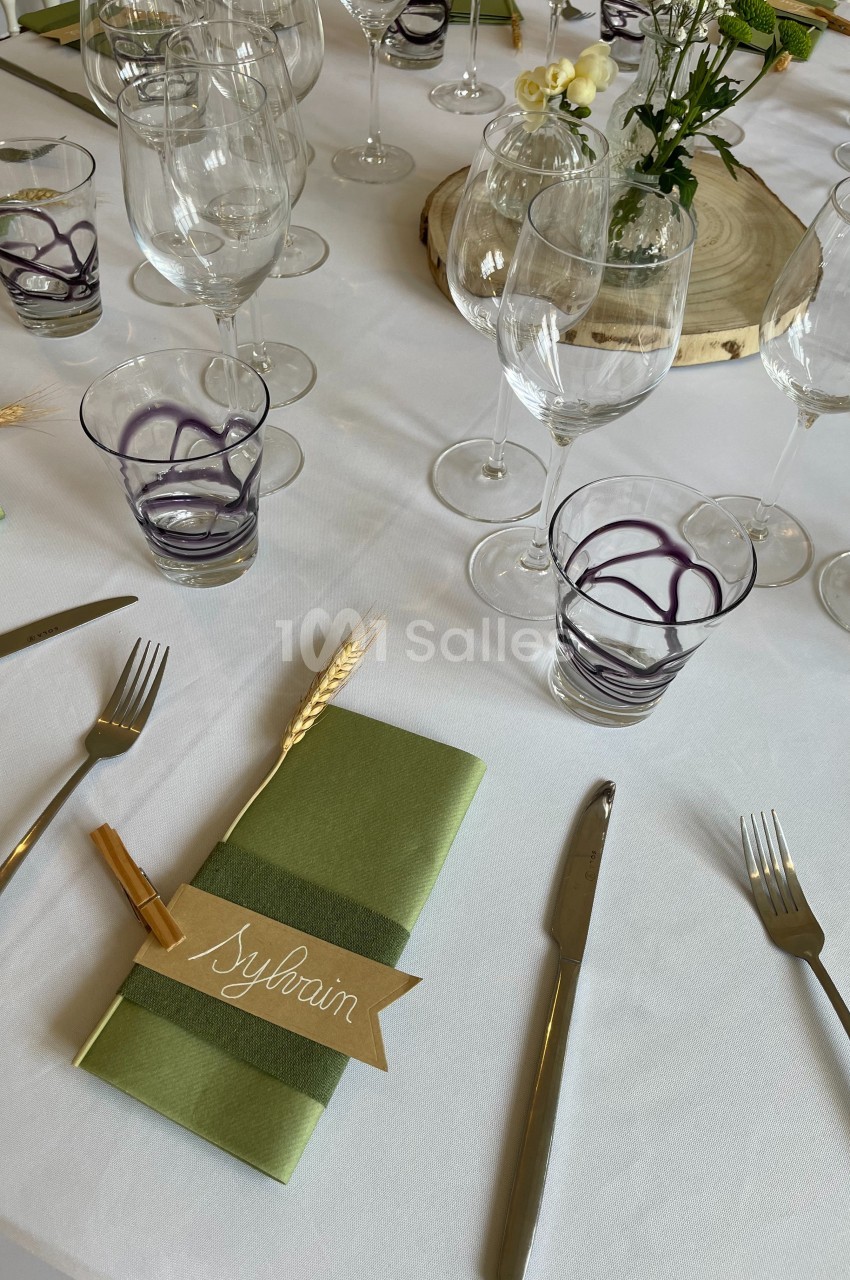 Table dressée avec des verres, couverts, serviettes vertes et marque-places personnalisés sur une nappe blanche.