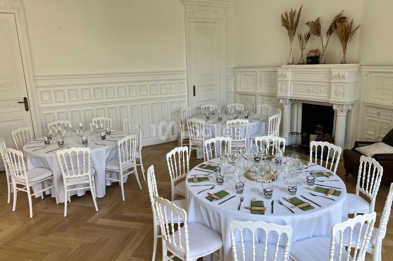 Salle élégante avec tables rondes dressées, chaises blanches, parquet clair et cheminée décorative en arrière-plan.