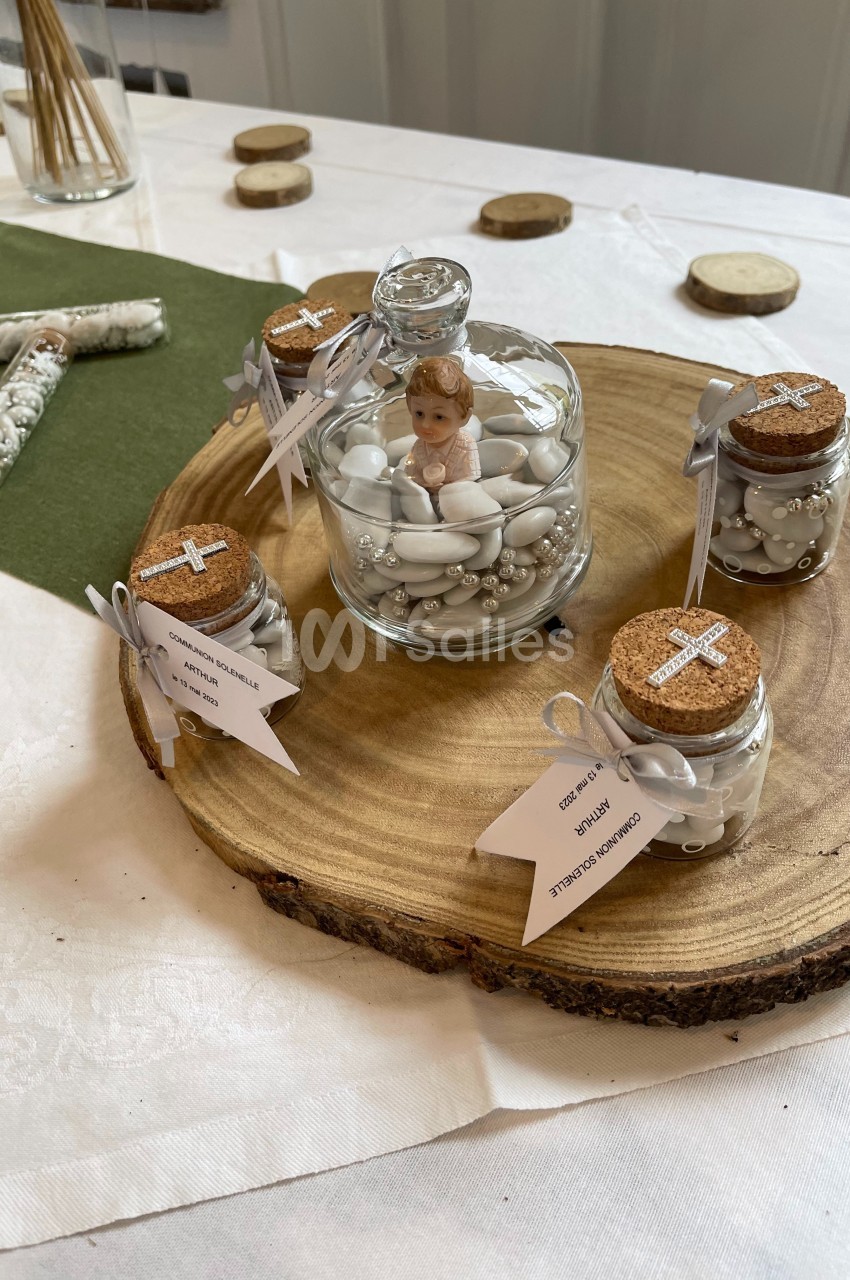 Petits pots de dragées et figurine sous cloche en décoration sur une rondelle de bois, disposés sur une table.