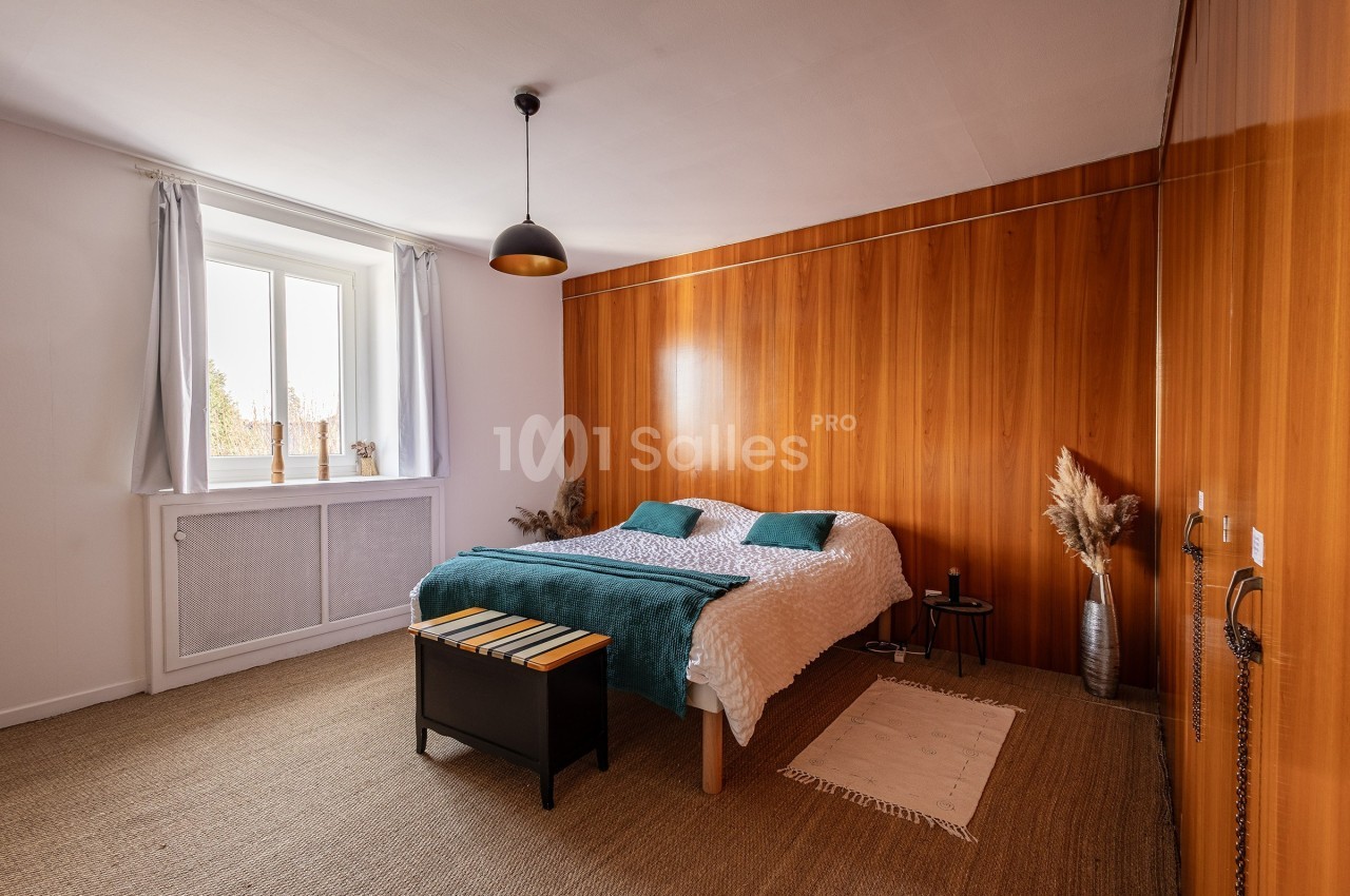 Chambre lumineuse avec lit double, mur en bois, tapis en jute, fenêtre avec rideaux blancs et décoration minimaliste.