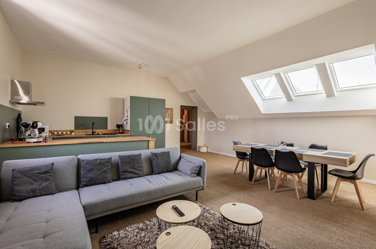 Salon lumineux avec canapé d'angle, table à manger, cuisine ouverte et fenêtres de toit laissant entrer la lumière naturelle.