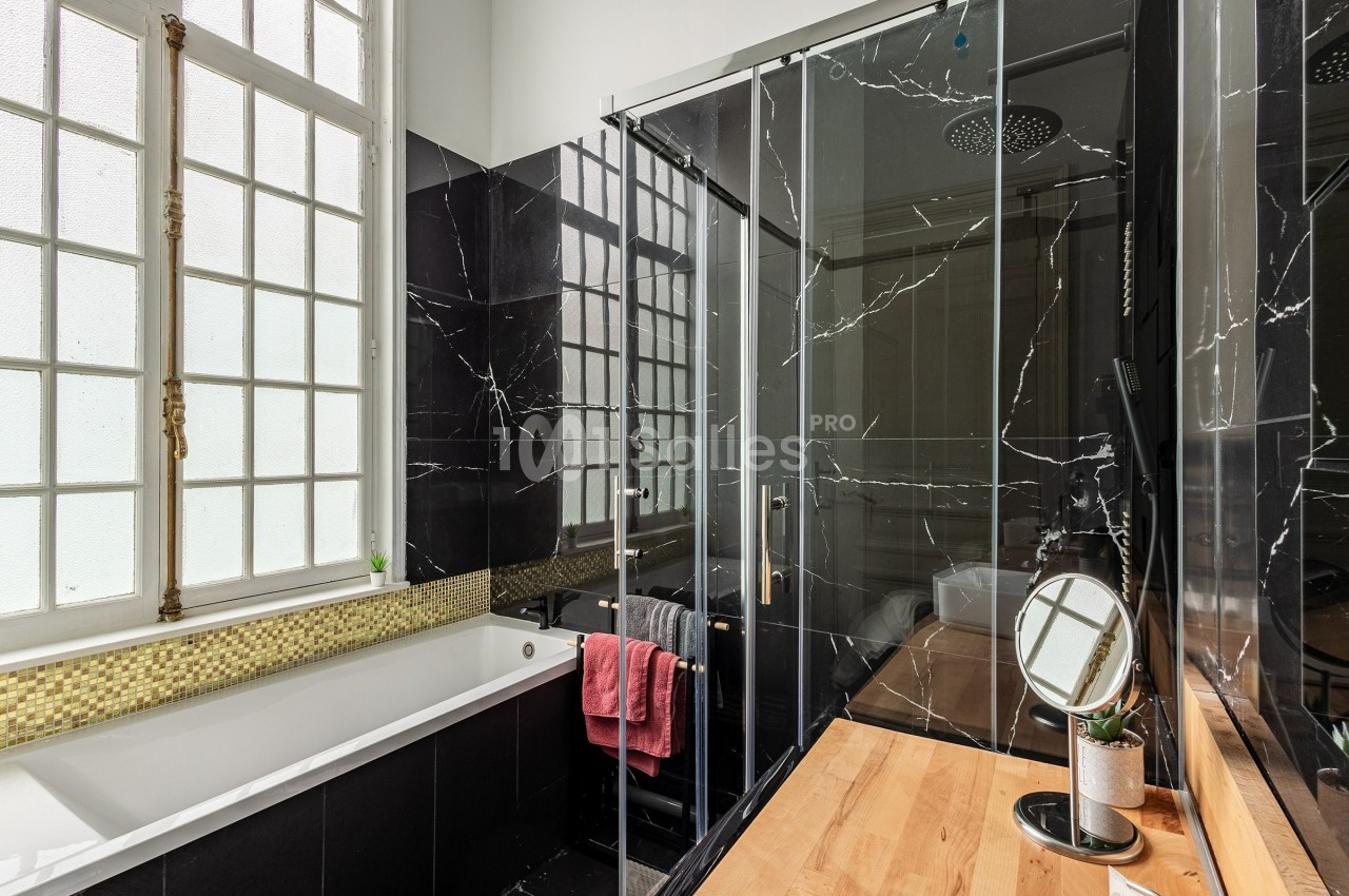 Salle de bain moderne avec baignoire, douche vitrée, murs noirs effet marbre, fenêtre à carreaux et meuble en bois clair.