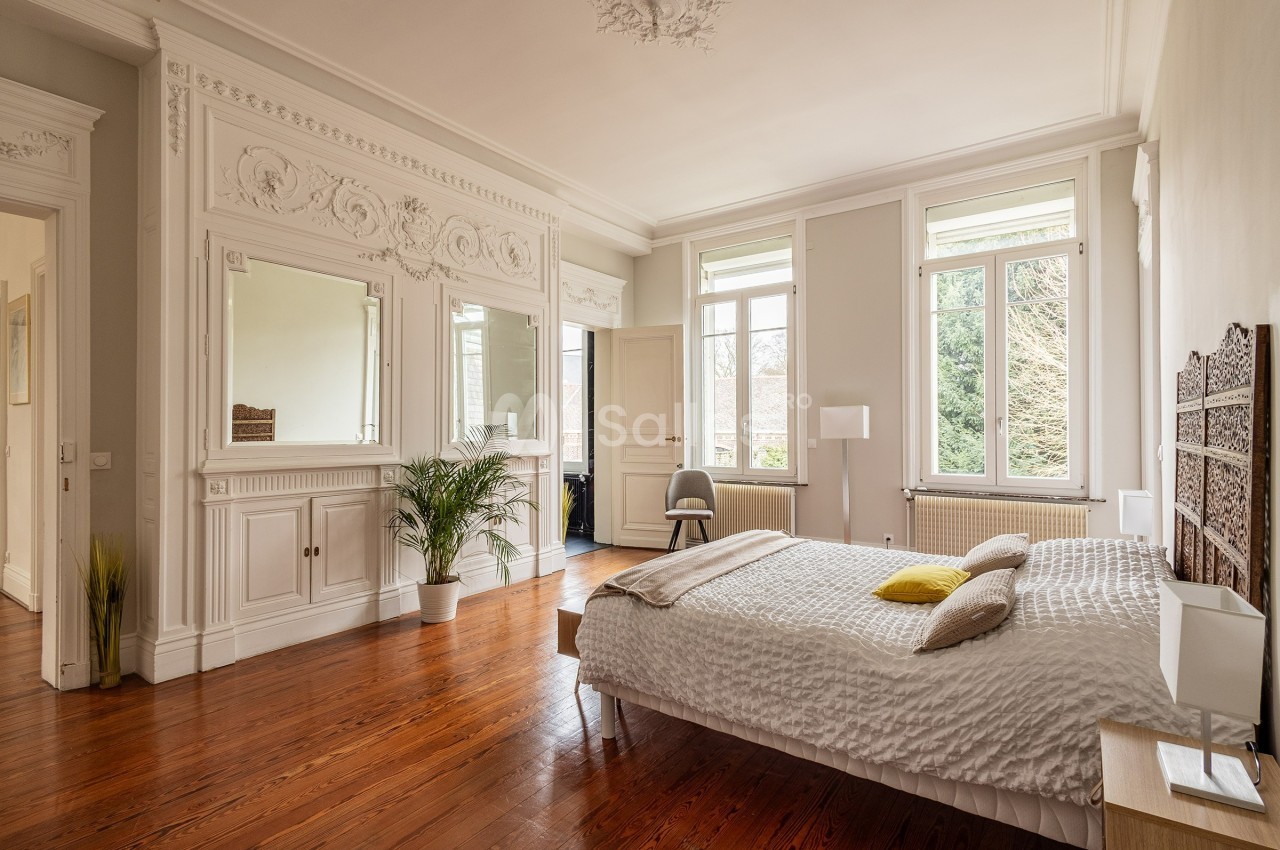 Chambre lumineuse avec parquet, lit double, moulures décoratives, miroirs et grandes fenêtres donnant sur un jardin.